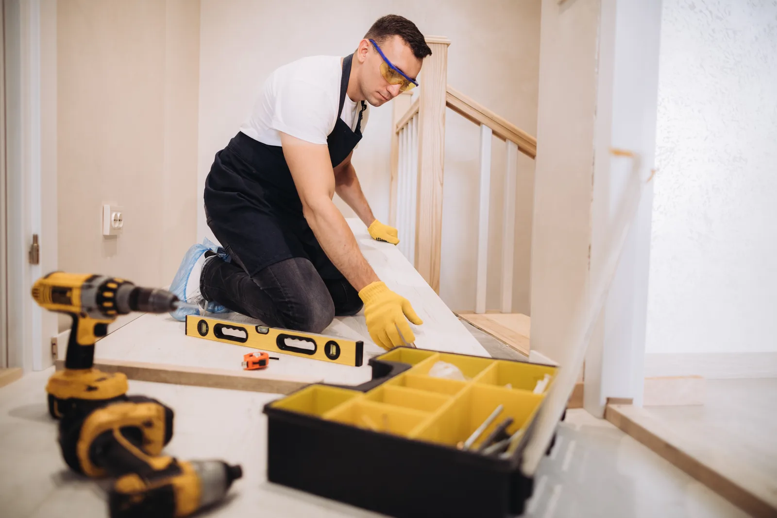 A craftsman in safety glasses fitting new flooring at the top of a staircase, with a level and tools beside him